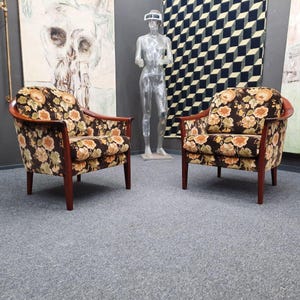 Könnte beinhalten: Zwei Vintage-Sessel mit dunklen Holzrahmen und floral gemusterten Polstern. Die Stühle haben eine geschwungene Rückenlehne und Armlehnen und stehen vor Kunstwerken und einem Wandbehang mit geometrischem Muster. Die Stühle stehen auf einem grauen Teppich.