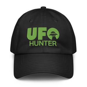 Può includere: Un cappellino da baseball nero con la scritta UFO HUNTER ricamata in verde brillante. La lettera O di UFO è sostituita da un grafico circolare che raffigura un disco volante che sorvola un raggio di luce, centrato sulla parte anteriore.
