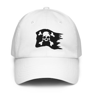 Pode incluir: Um boné de beisebol branco com um gráfico bordado em preto de uma bandeira pirata esfarrapada. O design exibe uma caveira com ossos cruzados no centro, com as bordas da bandeira parecendo desgastadas e desfiadas no lado direito.