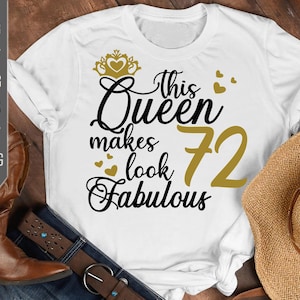 Puede incluir: Camiseta blanca con el texto "This Queen makes 72 look Fabulous" en negro y dorado. El diseño incluye una corona y corazones dorados. La camiseta se muestra con un par de botas de vaquero marrones, un sombrero y unos vaqueros.
