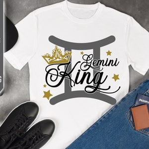 May include: White t-shirt with a gold crown and the text "Gemini King" in black lettering. The text is surrounded by a gray Gemini zodiac symbol. There are gold stars scattered around the design.