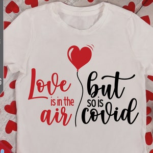 May include: White t-shirt with black text that reads "Love is in the air but so is covid". A red heart-shaped balloon is above the text.
