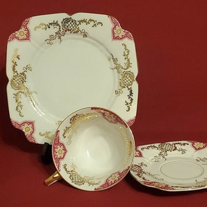 May include: A vintage tea set with a white background, featuring a square plate, a teacup, and a saucer. The set is decorated with gold filigree and pink floral accents. The teacup has a gold handle.