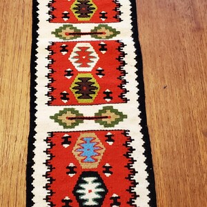 May include: A rectangular, woven table runner with a red, white, black, green, and blue geometric pattern. The runner has a black border and white fringe at both ends. The design features repeating shapes.