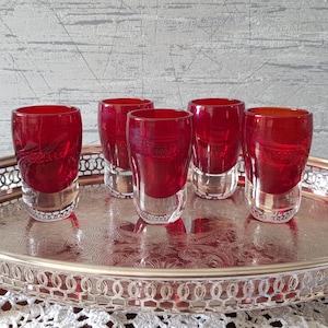 Puede incluir: Seis vasos de chupito rojos con bases transparentes están colocados en una bandeja plateada. Los vasos tienen un tono rojo vibrante y una forma redondeada. La bandeja tiene un borde decorativo y un patrón floral.