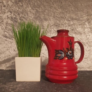 Puede incluir: Una tetera de cerámica roja con un diseño floral y un asa. La tetera está al lado de una maceta cuadrada blanca con hierba verde. La tetera tiene un borde dorado y un pico.