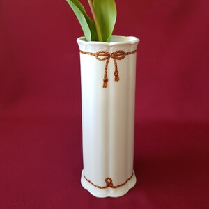 Op de afbeelding: Een witte keramische vaas met een gele tulp. De vaas heeft een geschulpte rand en is versierd met bruine, touwachtige strikken. De tulp heeft een heldergele bloem en groene bladeren. De achtergrond is effen bordeauxrood.