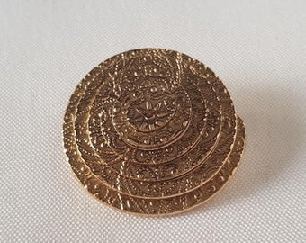 May include: A gold-colored, circular brooch with an intricate, layered design. The brooch features detailed patterns and textures, creating a vintage or antique aesthetic. The brooch is set against a white background.