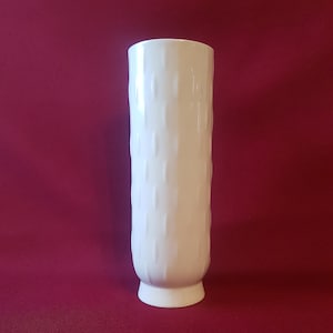 Puede incluir: Un jarrón alto de cerámica blanca con un patrón texturizado y repetitivo. El jarrón tiene forma cilíndrica con una base ligeramente ensanchada y un borde liso y redondeado. El fondo es de color rojo intenso.