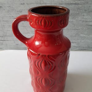 Puede incluir: Un jarrón de cerámica rojo brillante con asa. El jarrón tiene una base ancha, un cuello más estrecho y un borde acampanado. El cuerpo está decorado con un patrón repetido de formas florales estilizadas. Aproximadamente 20 cm de altura.