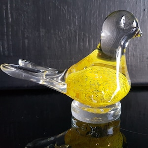 Może przedstawiać: Przezroczysta szklana figurka ptaka z żółtym korpusem i przezroczystymi skrzydłami i głową. Żółty korpus ma nakrapiany wzór. Ptak siedzi na przezroczystej, okrągłej podstawie. Figurka jest na ciemnym tle.