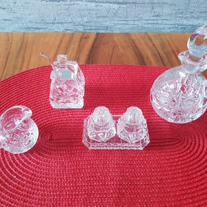 May include: A collection of cut crystal serving pieces, including a creamer, sugar bowl with spoon, salt and pepper shakers, and a small cruet with a handle and stopper. The pieces are arranged on a red woven placemat.