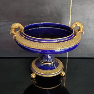 May include: A cobalt blue and gold decorative bowl with a pedestal base. The bowl has a wide, open top and is adorned with gold accents, including handles shaped like bird heads. The base features a gold trim and small feet.