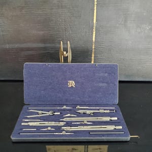 Può includere: Un set da disegno vintage in una custodia di velluto blu. Il set include diversi compassi, divisori e altri strumenti da disegno argentati. La custodia è aperta, rivelando gli strumenti ordinatamente disposti all'interno. Lo sfondo è una superficie scura e strutturata.