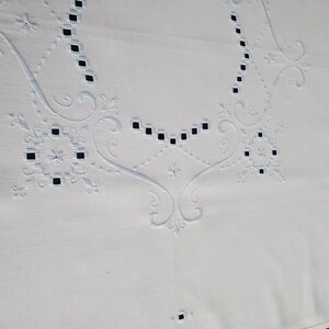 Peut inclure: Gros plan sur une nappe blanche avec des broderies complexes. Le motif présente des motifs floraux bleu clair, des formes géométriques et des carrés ajourés. La broderie délicate ajoute une touche d'élégance au textile.