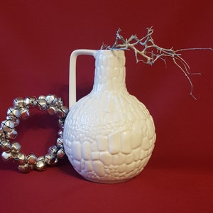 Könnte beinhalten: Eine weiße Keramikvase mit strukturierter Oberfläche und Henkel, neben einem silbernen Glockenkranz und einem silberfarbenen Ast, alles vor rotem Hintergrund. Die Vase hat eine bauchige Form.