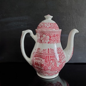 May include: A white and red ceramic teapot with a lid. The teapot features a red floral pattern on the lid and a red transferware scene of people and buildings. The handle and spout are white.