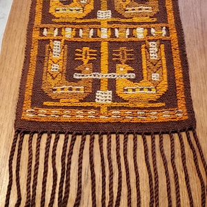 Puede incluir: Textil tejido marrón y naranja con un patrón geométrico repetitivo. El diseño presenta figuras y formas estilizadas en tonos naranja, amarillo y blanco sobre un fondo marrón. El textil tiene una franja de cordones marrones con detalles en naranja.
