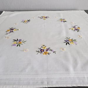Peut inclure: Nappe blanche avec broderie florale. La broderie présente des bouquets de fleurs jaunes, violettes et oranges avec des feuilles vertes, disposés en cercle. De petites fleurs blanches en forme de marguerite sont dispersées sur les bords.