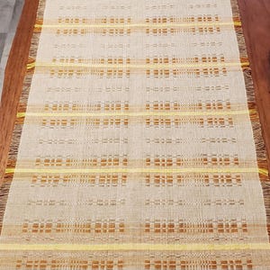 May include: Beige and yellow woven table runner with a geometric pattern. The runner has a rectangular shape with fringed edges. The design features horizontal stripes and blocks of color, creating a textured appearance. The runner is displayed on a wooden table.