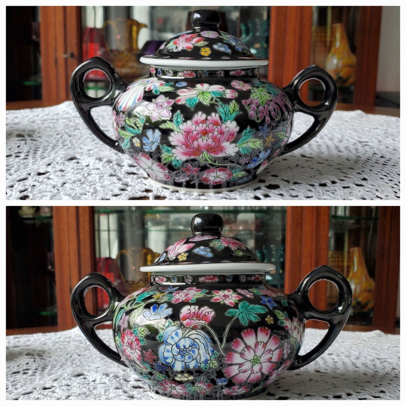 May include: A black ceramic sugar bowl with a lid, adorned with a vibrant floral pattern in pink, blue, and green. The bowl has two black handles and a black knob on the lid. The design is set against a black background.