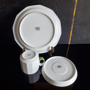 Op de afbeelding: Een wit porseleinen theeservies met een groen handvat. De set bevat een bord, een schotel en een kopje. Het bord en de schotel hebben een achthoekige vorm. De beker heeft een geometrisch ontwerp. Gemaakt in Duitsland.