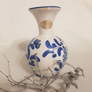 Puede incluir: Un jarrón de cerámica blanco con diseños florales azules y un borde azul. Una etiqueta dorada está adherida al jarrón. Una rama plateada está al lado del jarrón. El jarrón mide aproximadamente 15 cm de alto.