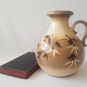 Puede incluir: Jarra de cerámica beige y marrón con asa, adornada con un diseño floral. Junto a ella, un libro antiguo con cubierta negra y bordes rojos. La jarra tiene una forma redondeada.