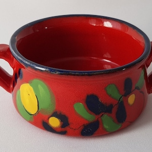 May include: A vibrant red ceramic bowl with two handles, adorned with hand-painted floral designs in green, yellow, and blue. The rim is accented with a thin blue line, creating a colorful and decorative piece.