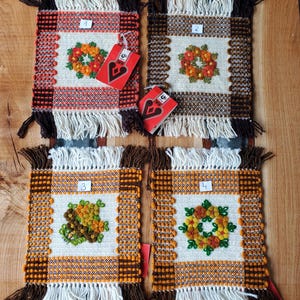 May include: A set of four square woven coasters with fringed edges. Each coaster features an embroidered floral design in orange, yellow, and green. The coasters have a woven border in brown, orange, and white.