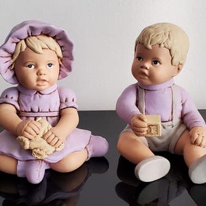 Könnte beinhalten: Zwei Babyfiguren aus Keramik. Eine trägt ein lavendelfarbenes Kleid und eine Haube und hält ein Spielzeug. Die andere trägt ein lavendelfarbenes Hemd, Hosenträger und Shorts und hält einen Block. Beide haben blondes Haar und sitzen.