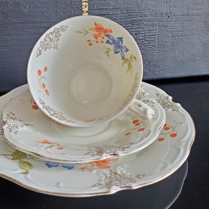 May include: A cream-colored porcelain teacup and saucer set featuring a delicate floral pattern with orange and blue flowers, green leaves, and small red berries. The edges are decorated with ornate gold filigree and scalloped rims.