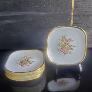 May include: A set of white square saucers with gold rims. Each saucer features a floral design with pink and yellow flowers. The saucers are stacked and displayed on a black surface.
