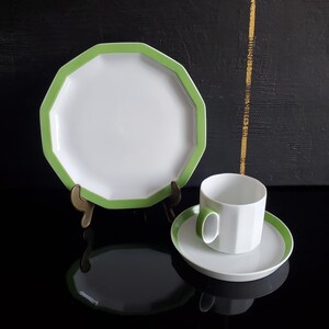 Op de afbeelding: Witte achthoekige bord met groene rand, gepresenteerd op een messing standaard. Een bijpassende kop en schotel met groene accenten zijn ook aanwezig. De set staat op een reflecterend zwart oppervlak.