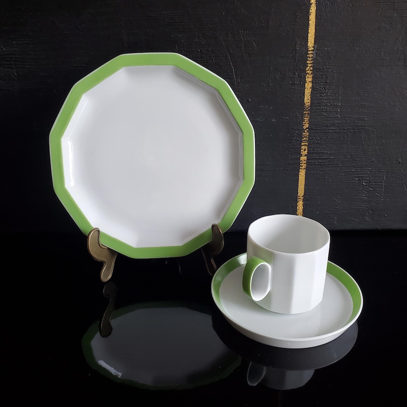 Op de afbeelding: Witte achthoekige bord met groene rand, gepresenteerd op een messing standaard. Een bijpassende kop en schotel met groene accenten zijn ook aanwezig. De set staat op een reflecterend zwart oppervlak.