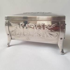 Peut inclure: Une boîte à bijoux rectangulaire argentée avec des détails ornés. La boîte présente un relief décoratif de deux figures ailées encadrant un emblème central. Elle a des côtés incurvés et repose sur quatre petits pieds incurvés. Le couvercle a un dessus légèrement incurvé.