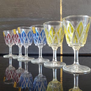 Puede incluir: Un conjunto de seis copas de licor vintage. Las copas presentan cuencos transparentes con diseños de diamantes en rojo, azul y amarillo. Cada copa tiene un tallo y una base transparentes, que reflejan la luz. Las copas están dispuestas en fila, mostrando sus coloridos diseños.