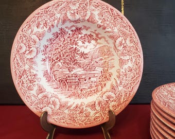 May include: A round ceramic bowl featuring a red and white transferware pattern. The center depicts a rural landscape with a horse-drawn cart, trees, and sheep. The wide rim is decorated with a dense floral border of blossoms and leaves.