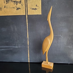 May include: A wooden sculpture of a crane with a long, curved neck and slender legs. The bird is light brown and stands on a rectangular base. The background features a dark blue wall with gold accents.