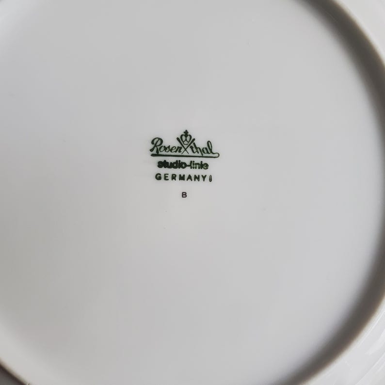 Op de afbeelding: Een witte porseleinen bord met een gebogen rand. Het bord heeft een groen logo met de tekst "Rosenthal studio-linie GERMANY" en de letter "B". Het bord is schoon en lijkt ongebruikt.