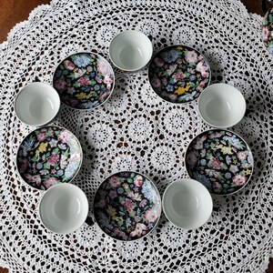 May include: A set of eight pieces of fine china, including four small white bowls and four matching saucers. The saucers feature a black background with a colorful floral design in pink, blue, yellow, and green. The set is arranged on a white lace tablecloth.