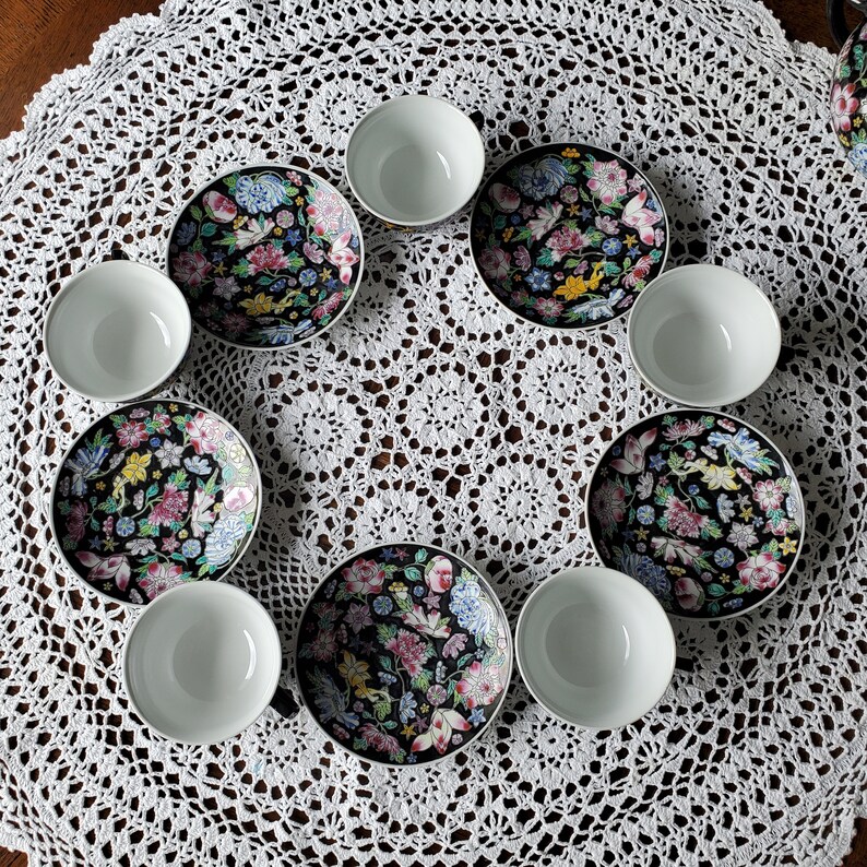 May include: A set of eight pieces of fine china, including four small white bowls and four matching saucers. The saucers feature a black background with a colorful floral design in pink, blue, yellow, and green. The set is arranged on a white lace tablecloth.
