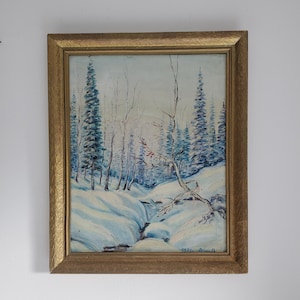 May include: A framed landscape painting depicting a snowy forest scene. The artwork features a stream winding through snow-covered banks, with evergreen trees and bare trees under a pale blue sky. The frame is gold-colored.