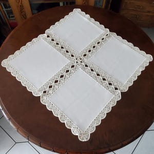 Op de afbeelding: Vier witte vierkante placemats met een crèmekleurige, opengewerkte kanten rand en geschulpte randen. De placemats zijn gerangschikt op een donkerbruine, ronde houten tafel. De kanten rand creëert een decoratief patroon.