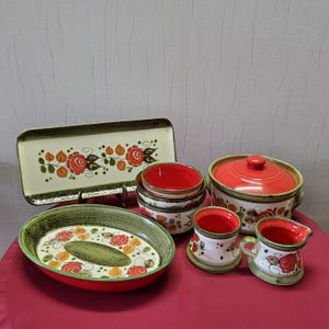 Könnte beinhalten: Eine Sammlung von Keramik-Servierschalen mit floralem Design. Das Set umfasst ein rechteckiges Tablett, eine ovale Schale, mehrere Schalen, einen Topf mit Deckel und zwei kleine Krüge. Die Teile weisen rote, orangefarbene und grüne Blumenmuster auf cremefarbenem Hintergrund auf.