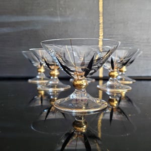 May include: Set of six clear glass dessert or cocktail glasses. Each glass features a gold-colored base and rim, with black and gold decorative accents. The glasses are arranged on a reflective black surface.