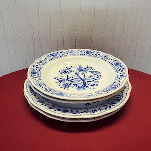 May include: Three white ceramic plates stacked on top of each other, each with a blue floral pattern. The plates have a scalloped edge and a detailed blue design around the rim and in the centre. The design features a stylized tree and other floral elements.