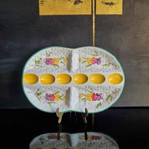 May include: Oval-shaped serving dish with a white woven pattern, green trim, and floral accents. The dish has six yellow egg-shaped indentations. The dish is displayed on a gold-colored stand against a dark background.