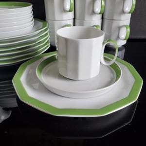 Op de afbeelding: Witte keramische servies met een groene rand en handvat. De set bevat borden, schotels en bekers. De borden hebben een achthoekige vorm. De bekers zijn op de achtergrond gestapeld.