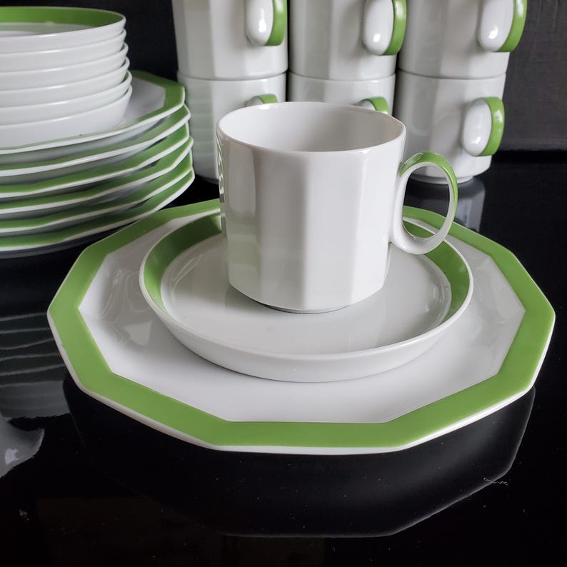 Op de afbeelding: Witte keramische servies met een groene rand en handvat. De set bevat borden, schotels en bekers. De borden hebben een achthoekige vorm. De bekers zijn op de achtergrond gestapeld.